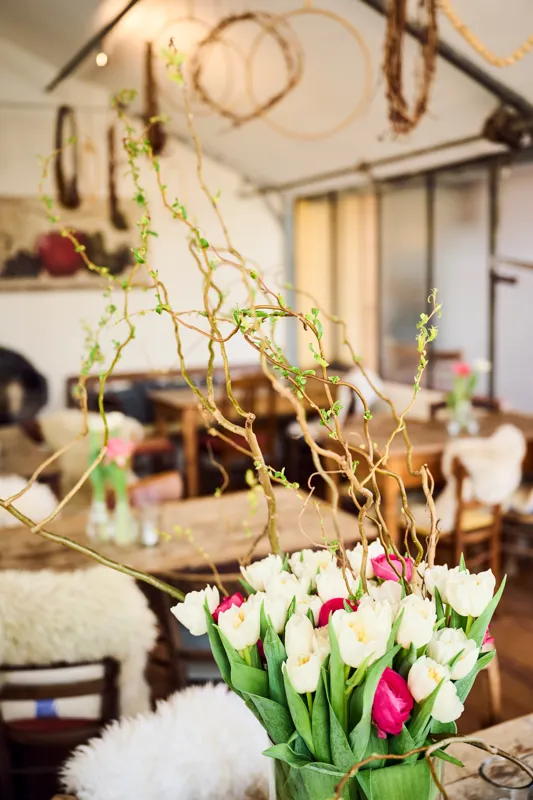 A decorative floral arrangement featuring white tulips with pink accents in a natural twig base, placed on a rustic surface. The background includes warm lighting and rustic wooden elements, enhancing a cozy, elegant setting.