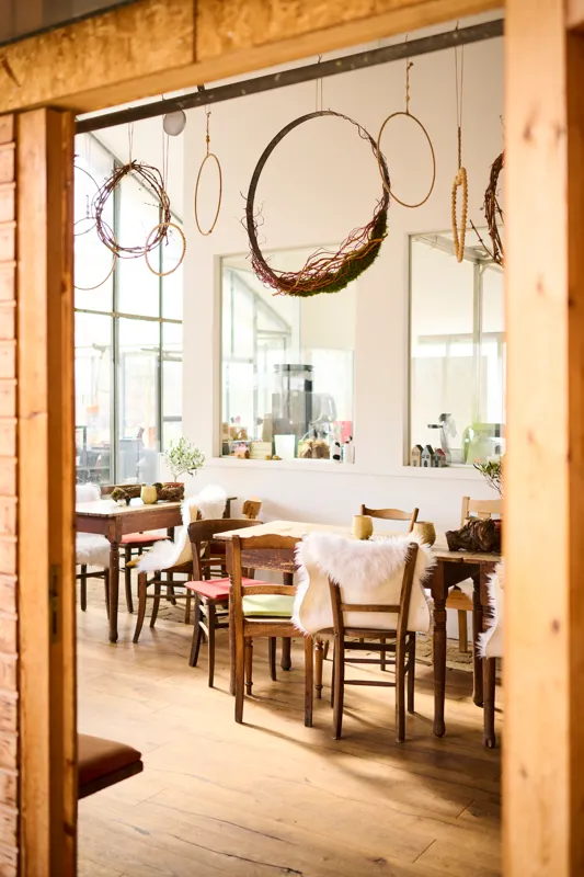 Elegant indoor dining area featuring rustic wooden furniture with plush fur throws on chairs, framed by large windows allowing natural light. Decorative wreaths and circular hanging ornaments add a cozy, artistic ambiance.