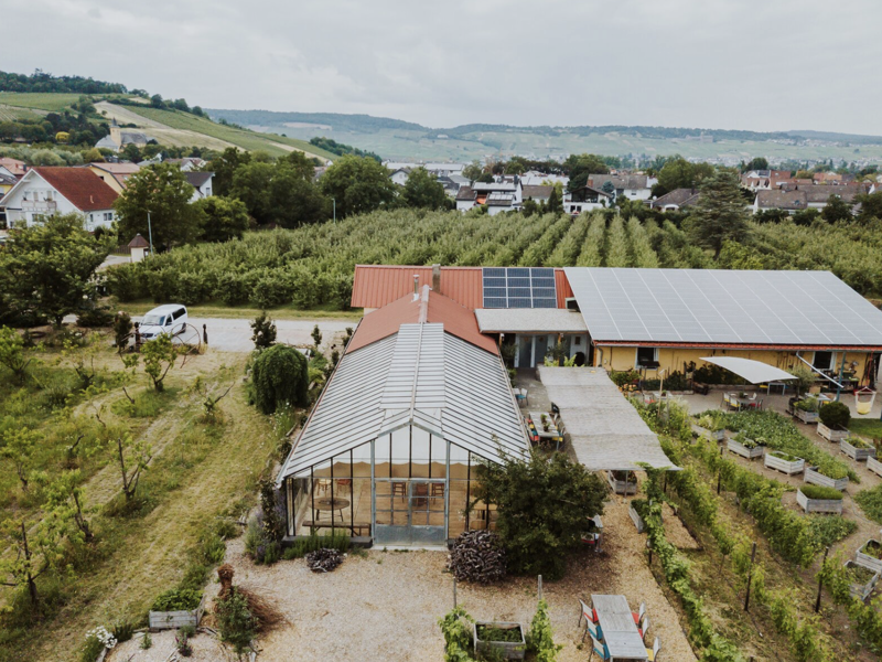Aerial view of a rural facility featuring modern agricultural structures, including a greenhouse with large glass panels and solar panels on adjacent buildings. The setting includes lush vineyards and residential houses in a scenic, hilly landscape.
