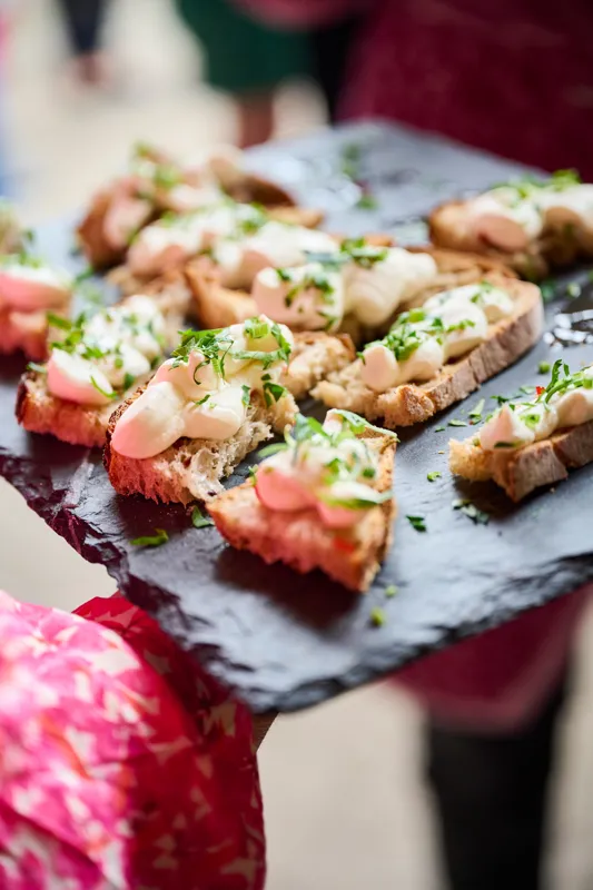 A neatly arranged charcuterie board featuring assorted small sandwiches with fresh toppings, including slices of ham, cheese, and garnished with herbs and dill. The presentation is on a textured surface, likely part of an outdoor or casual dining event setup.