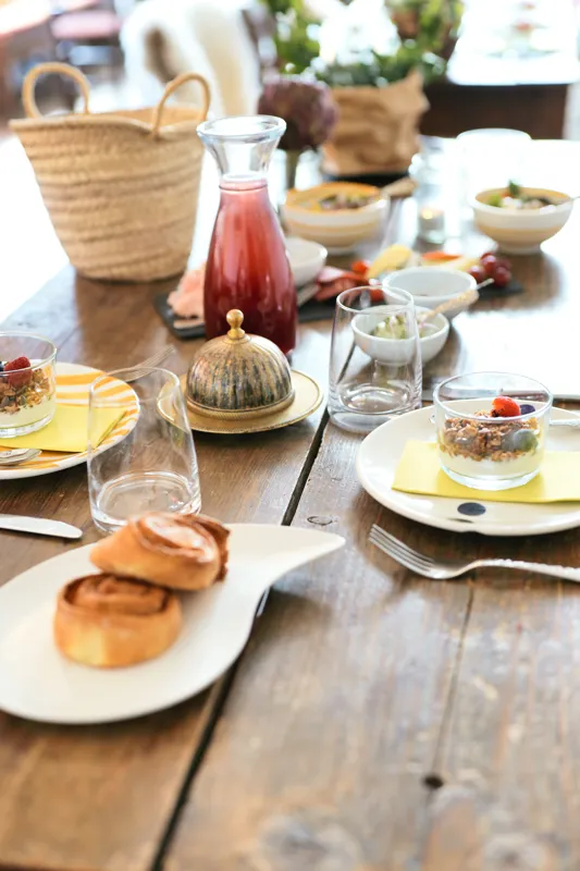 A meticulously arranged breakfast buffet features an assortment of small plates with savory and sweet dishes, including pastries, fruit, and creamy desserts. Glassware, decorative pitchers, and natural wooden tableware create a refined, inviting presentation.