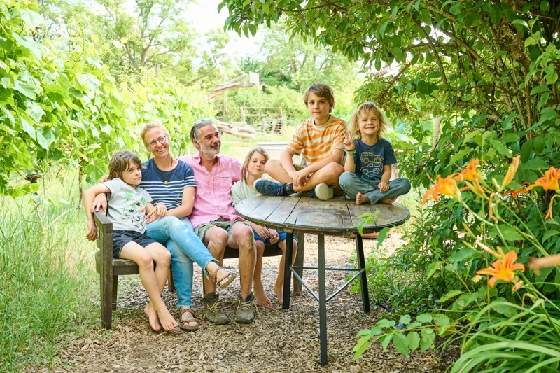 A family of five sits outdoors on a wooden bench and table surrounded by lush greenery. Bright orange flowers and natural lighting create a vibrant, relaxed garden setting.