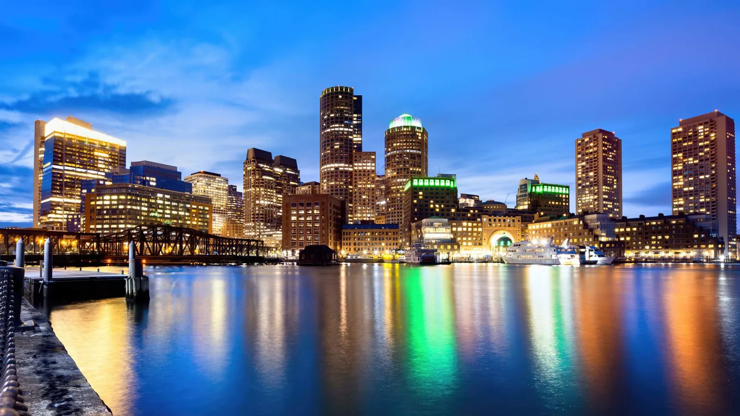 Modern cityscape featuring illuminated high-rise buildings along a waterfront during twilight. Reflective water displays vibrant lighting from the skyscrapers and nearby docked vessels.