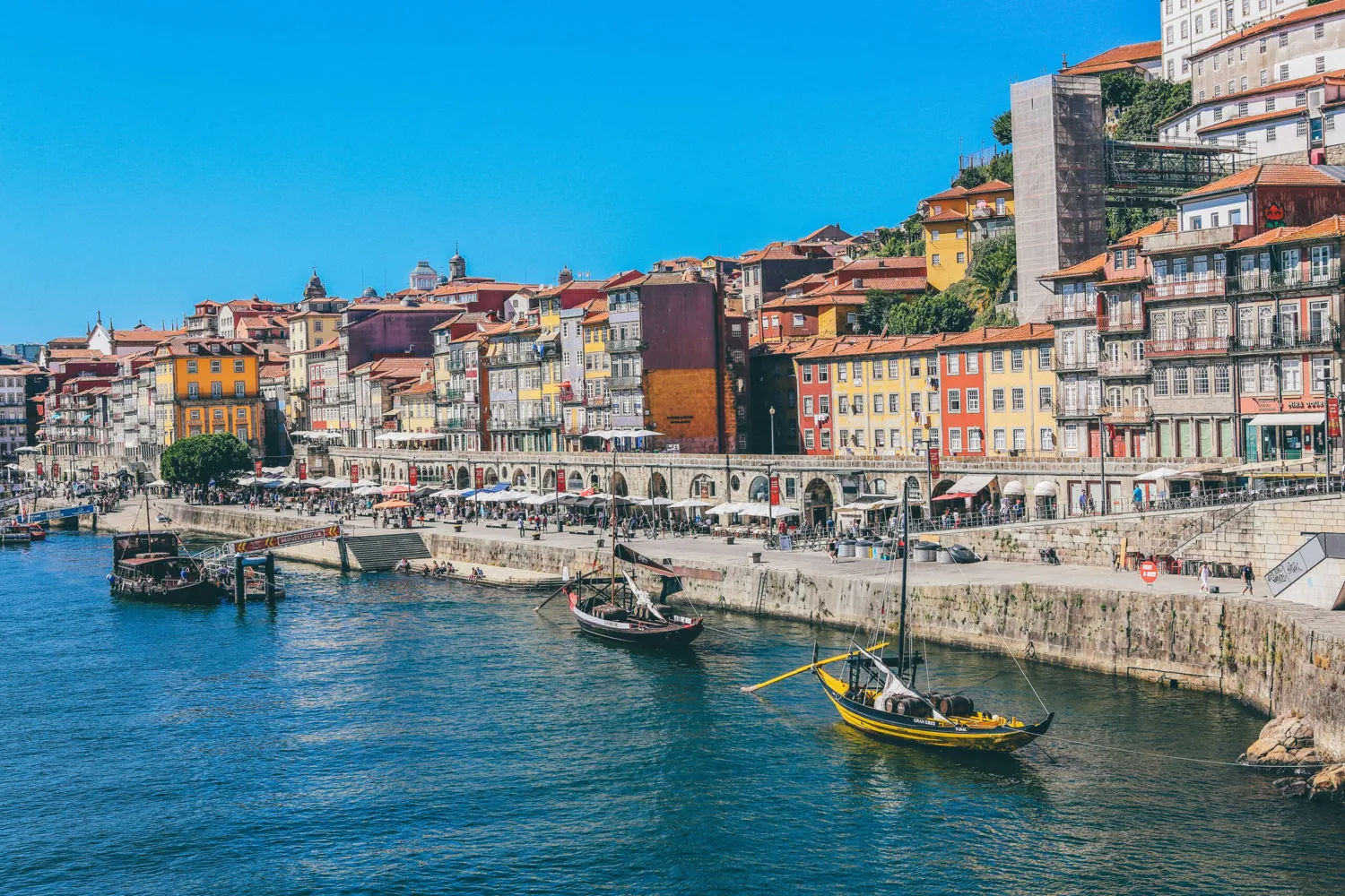 Brightly painted historic buildings line a vibrant harbor, featuring traditional boats docked along the waterfront promenade under clear skies. Outdoor seating and market stalls add to the lively atmosphere of this picturesque coastal setting.