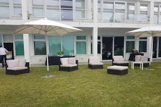 Modern outdoor seating arrangement featuring cushioned lounge chairs and circular benches under large patio umbrellas, set against a contemporary building facade with large windows. The area appears to be part of an event or relaxation space with well-maintained grass and greenery.