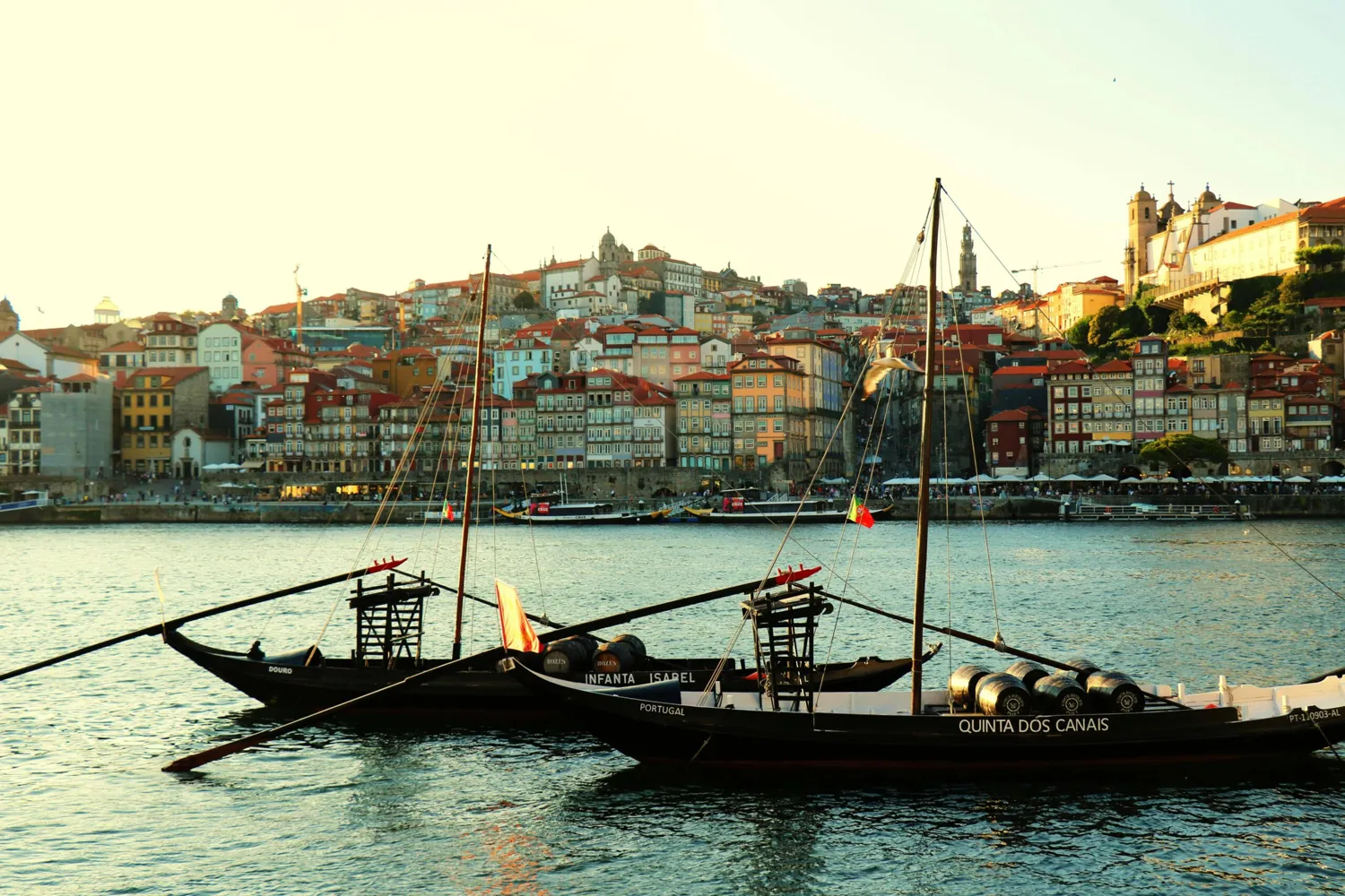 Historic wooden boats on the Douro River in front of colorful, historic buildings and a church tower in a scenic hilly landscape. Traditional craft with masts and flags reflects cultural heritage.