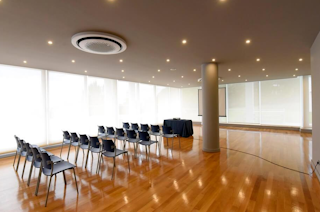 Spacious conference room featuring polished wooden flooring, modern seating arrangements with individual chairs, and large glass walls providing natural light. Central ceiling fixture and sleek design elements enhance the professional meeting space.