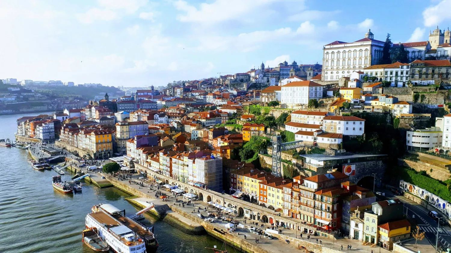 A picturesque aerial view of a historic European waterfront featuring colorful buildings with terracotta roofs along a riverbank. Prominent architectural landmarks and a bustling promenade with boats docked along the shoreline are visible.