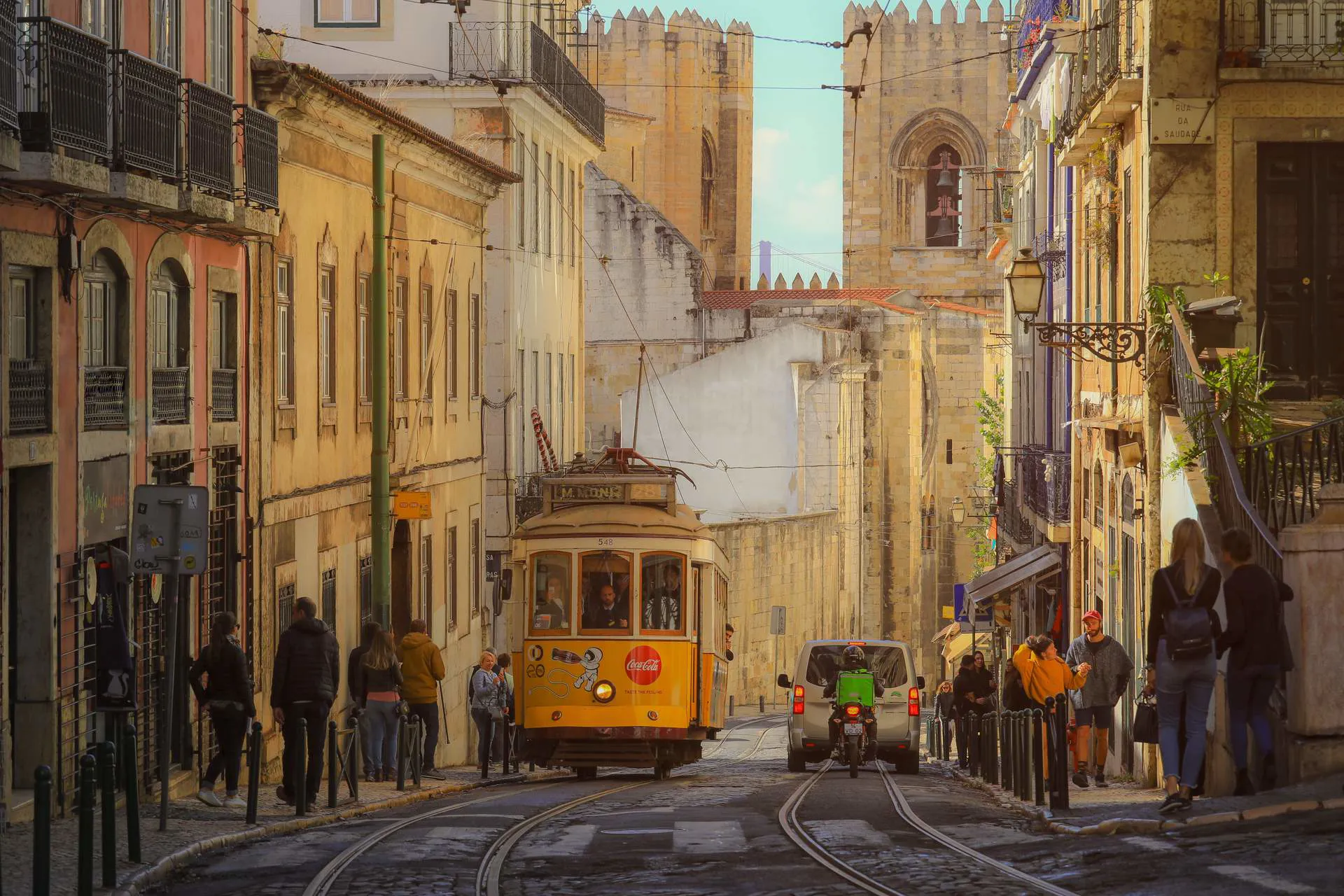 Historic yellow tram travelling through a narrow cobblestone street in Lisbon with pedestrians and traditional buildings