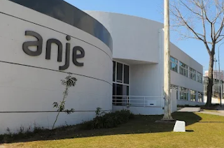 Modern architectural building with the "anje" logo prominently displayed on the curved facade. The structure features clean lines, a sleek design, and a well-maintained outdoor area with grass and minimalist landscaping.