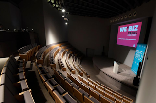 A spacious conference room with tiered seating facing a stage, illuminated by overhead lighting. The stage displays a presentation screen with branding for a business event.